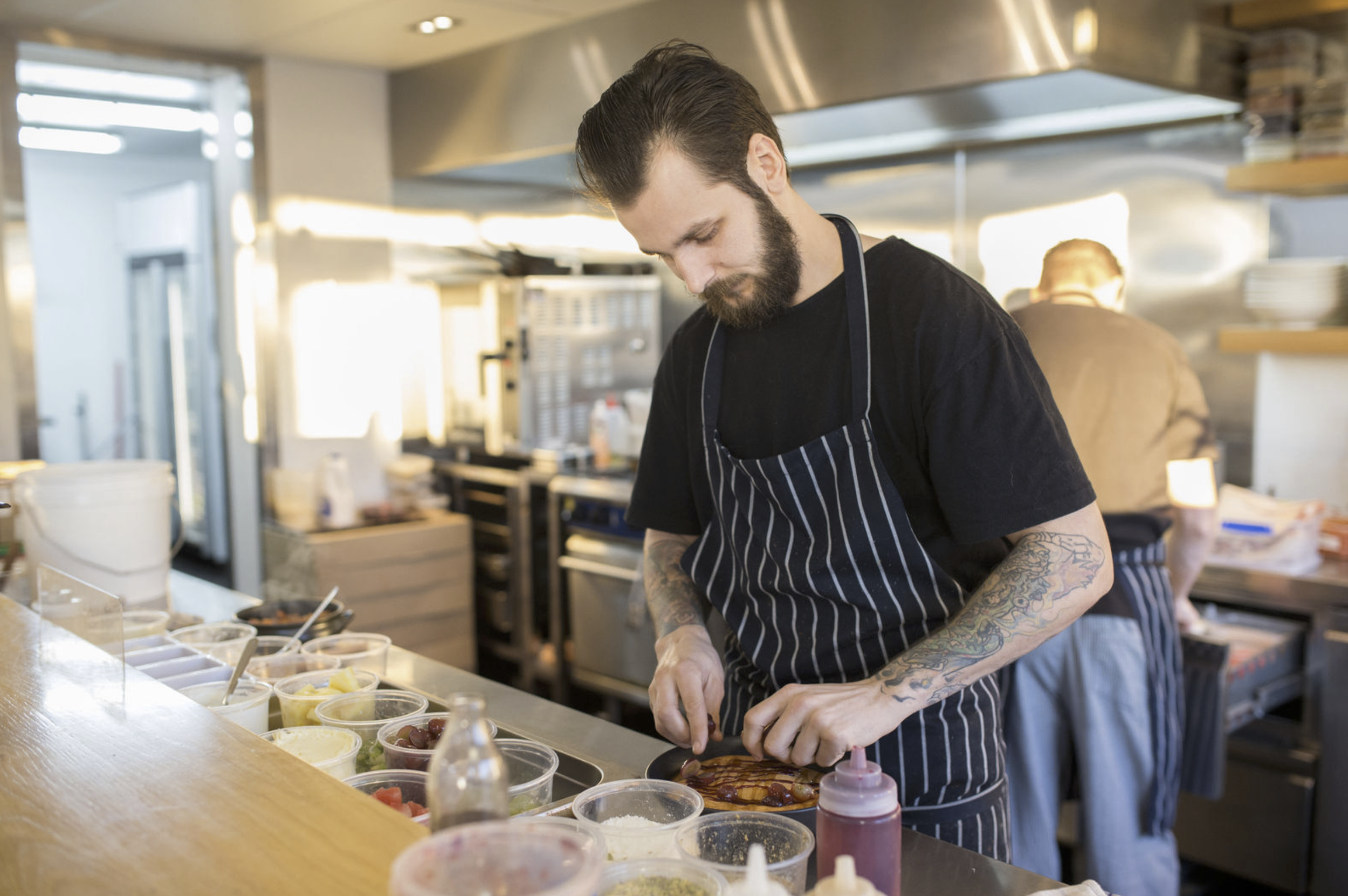 A male chef wearing a black shirt and striped apron decorating a dish during a commercial shoot